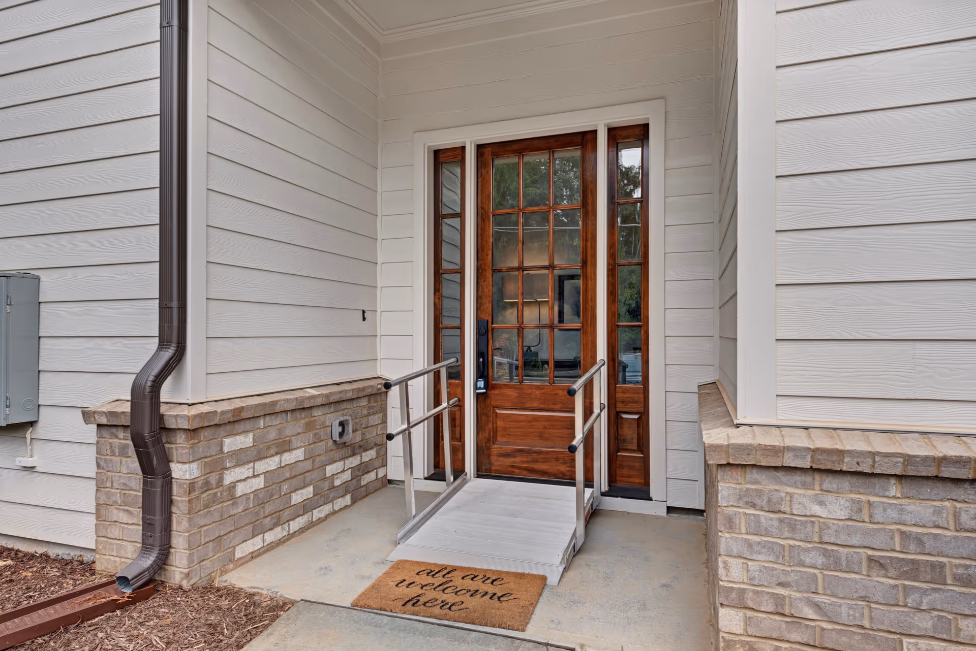 Entrance to a building with a wooden door featuring multiple glass panes, a small ramp with metal handrails, and a doormat that reads 'all are welcome here'. The exterior walls are light-colored with brick accents at the base.