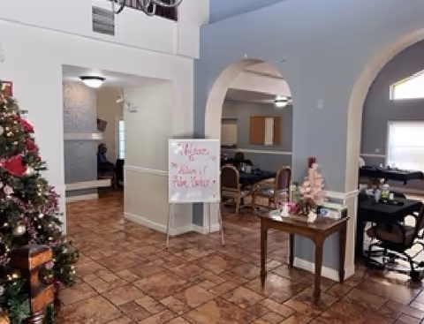 Interior view of a senior living facility common area with tiled floors, arched doorways, and a Christmas tree decorated with ornaments. There is a whiteboard sign in the center with red writing, several tables and chairs in the background, and a small table with holiday decorations on the right.