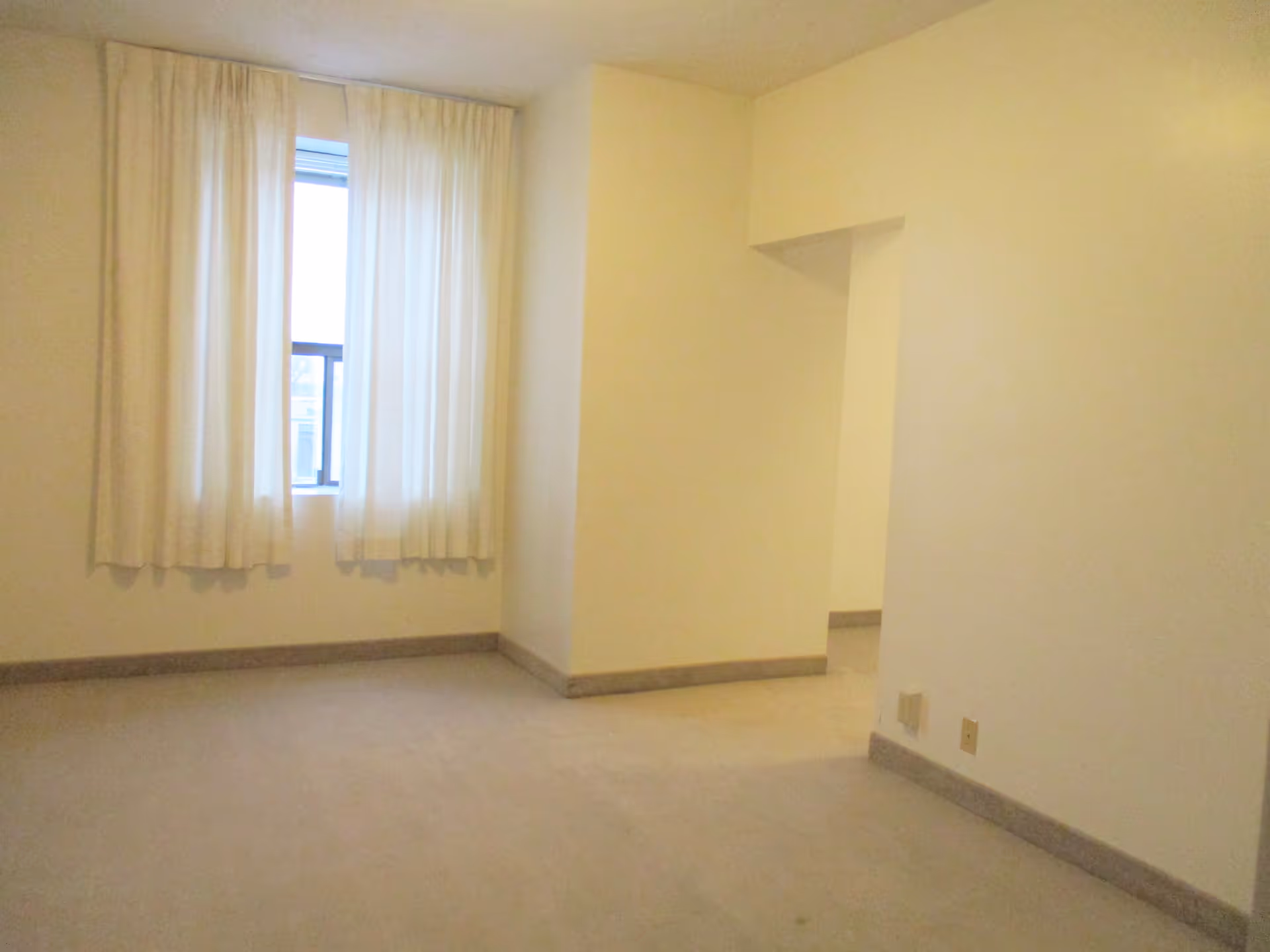 Empty room with beige carpet and cream-colored walls, featuring a window with white curtains and an open doorway leading to another room.
