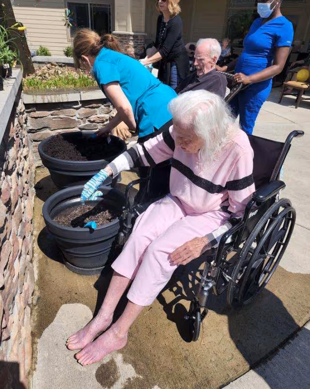 An elderly woman in a wheelchair plants soil in outdoor pots while caregivers assist in a courtyard.