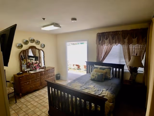 A cozy bedroom with a wooden bed frame, a neatly made bed with pillows, a wooden dresser with a large mirror, decorative plates on the wall, a lamp on a bedside table, and a window with brown curtains letting in natural light.