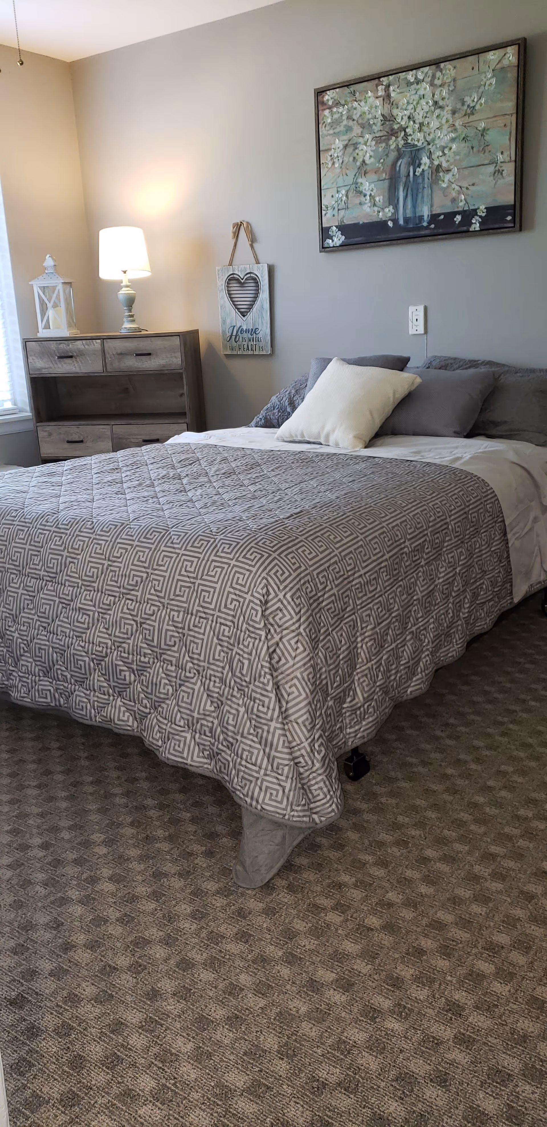 A cozy bedroom with a neatly made bed featuring a gray patterned quilt and multiple pillows. Next to the bed is a wooden dresser with a lamp and a decorative lantern on top. On the wall above the bed hangs a painting of white flowers in a blue vase, and a small hanging sign with a heart and the words 'Home is where the heart is'. The room has a carpeted floor and neutral-colored walls.