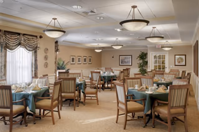 A dining room in a senior living facility with multiple round tables covered with green tablecloths, each set with napkins, glasses, and silverware. The room has beige walls, framed pictures, large windows with draped curtains, and several ceiling light fixtures. Wooden chairs with cushioned seats surround the tables.
