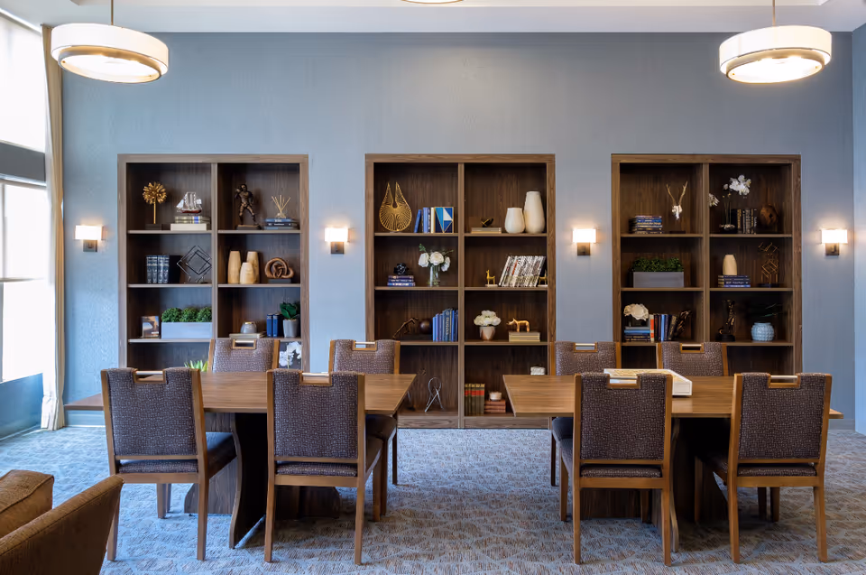 A bright communal dining room with wooden tables and chairs arranged in front of three built-in shelving units filled with books and decorative items.