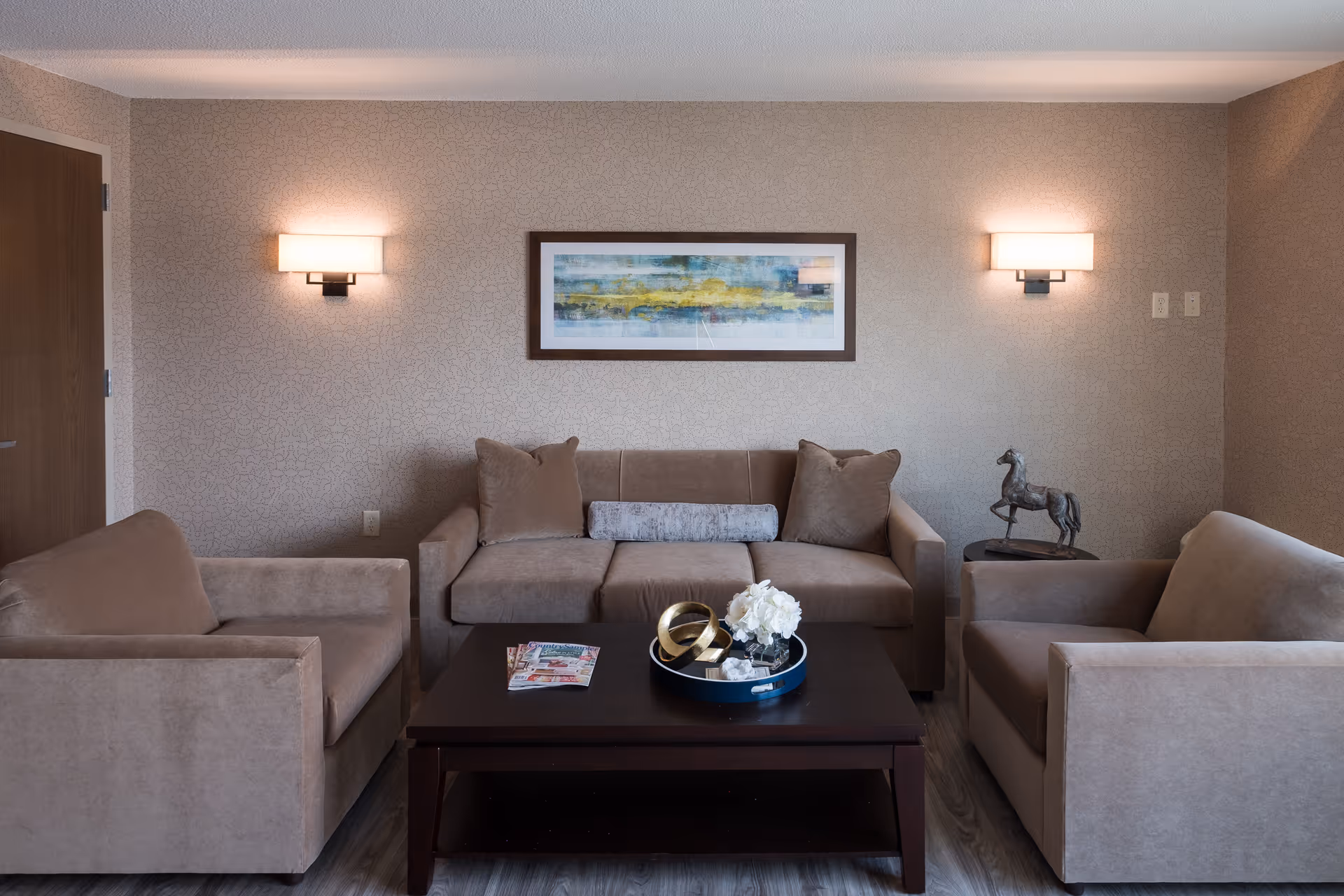 A cozy living room with a beige sofa and two matching armchairs arranged around a dark wooden coffee table. On the coffee table, there is a decorative tray with white flowers and golden rings, along with some magazines. The wall behind the sofa features a framed abstract painting and two wall-mounted lights. A small side table with a horse sculpture is placed next to one armchair.