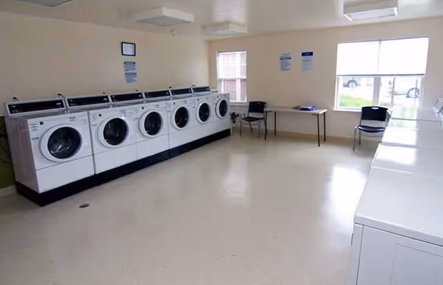 A laundry room with a row of front-loading washing machines on the left side and top-loading dryers on the right side. There are two windows letting in natural light, a table with some items on it, and two chairs near the windows. The room has a clean, simple design with light-colored walls and flooring.