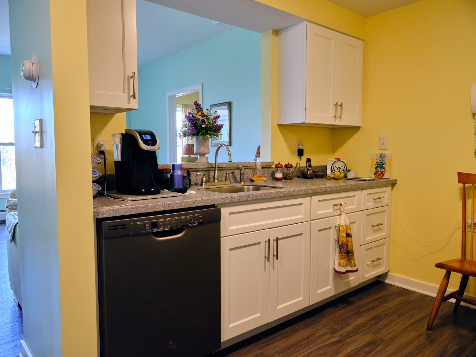 A kitchen area with white cabinets and drawers, a gray countertop, and a black dishwasher. On the countertop, there is a coffee maker, a purple cup, a vase with purple flowers, a phone, and some small containers. The walls are painted yellow, and there is a wooden chair to the right. A window and a room with light blue walls are visible in the background.