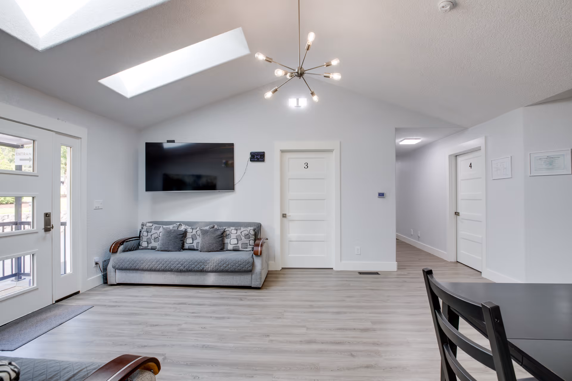 Bright communal living room with a sofa under a wall-mounted TV, numbered doors (3 and 4), skylights and a modern chandelier.