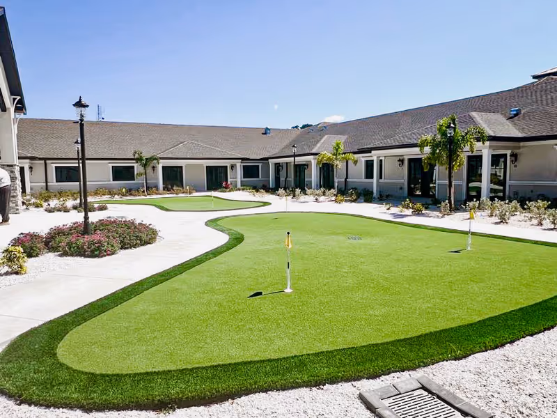 Outdoor courtyard area with a putting green surrounded by a paved walkway, small palm trees, and landscaping with flowers and shrubs. The courtyard is enclosed by a single-story building under a clear blue sky.