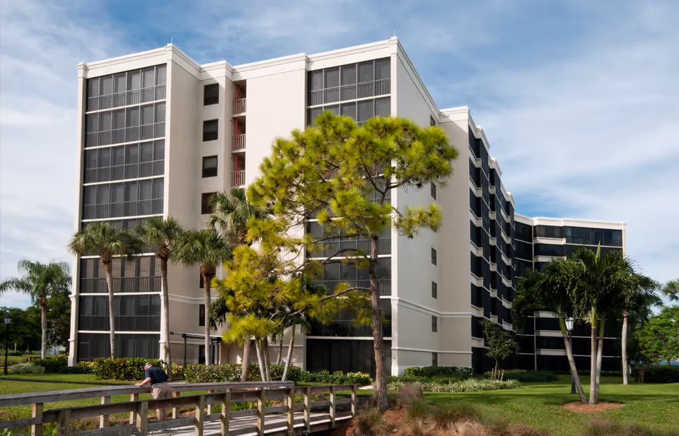 Exterior view of a multi-story senior living facility building with large screened balconies, surrounded by palm trees and greenery under a partly cloudy sky. A person is seen leaning on a wooden railing in the foreground.