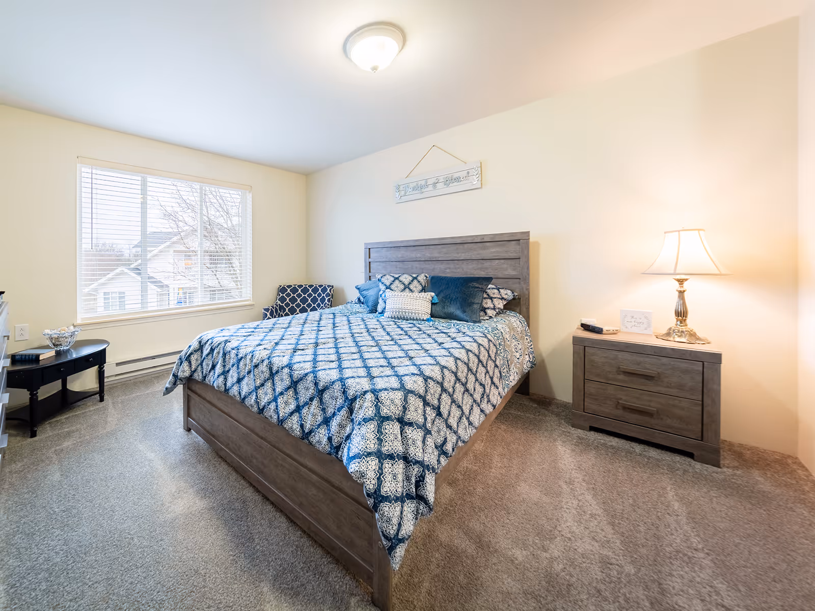 A bright bedroom with a large window on the left letting in natural light. The room features a wooden bed with a blue and white patterned bedspread and several pillows. To the right of the bed is a wooden nightstand with a lamp and a small decorative sign. There is a patterned armchair in the corner near the window and a small black table with decorative items on the left side of the room. The walls are painted a light cream color and the floor is carpeted.
