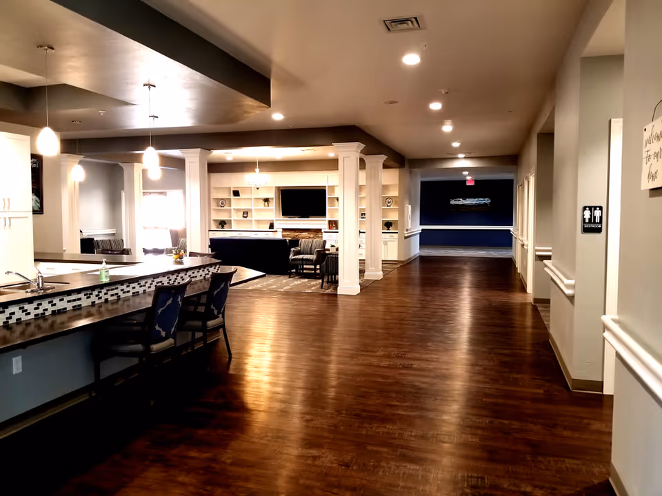 Interior view of a senior living facility showing a long hallway with wooden flooring, a kitchen counter with bar stools on the left, and a living area with chairs and a TV in the background. The hallway has ceiling lights and a restroom sign on the right wall.