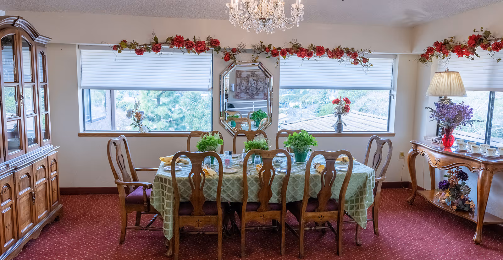 Sunlit dining room with a table set for six, wooden chairs and cabinets, floral decorations, and large windows overlooking trees.