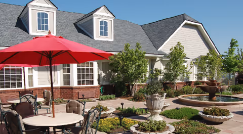 Sunlit courtyard with patio tables and a red umbrella, decorative planters and a fountain in front of a residential building.