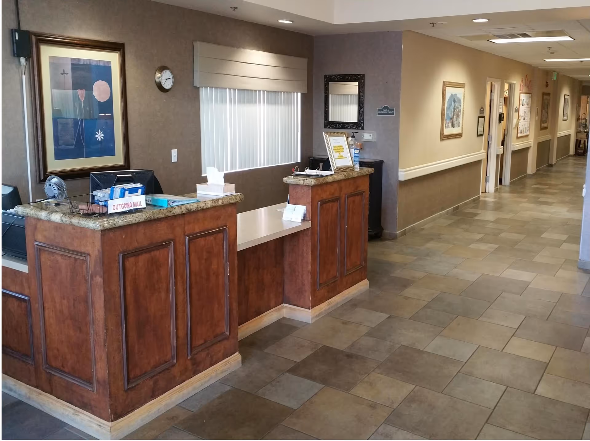 Reception desk area inside Chaparral Winds Assisted Living facility with a wooden counter, computer monitors, an outgoing mail basket, a clock on the wall, framed artwork, and a hallway extending to the right with several doors and framed pictures.