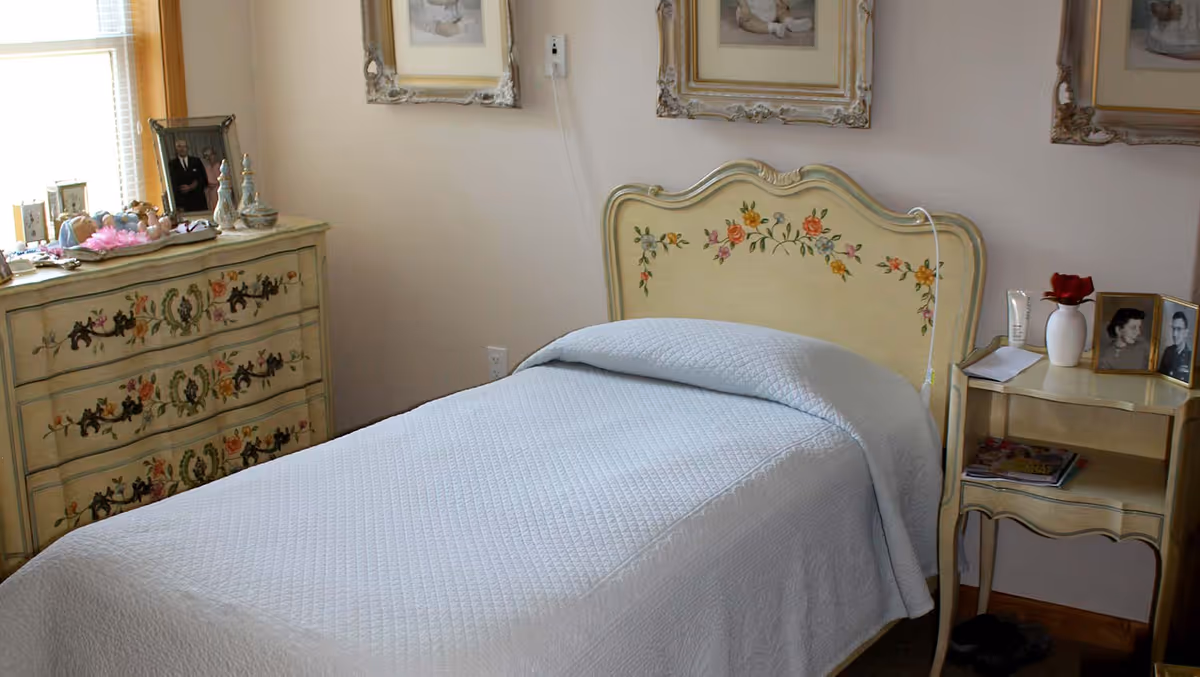 A neatly made single bed with a light blue quilted bedspread in a bedroom. The bed has a decorative headboard with floral designs. To the left of the bed is a dresser with floral patterns and various framed photos and decorative items on top. To the right of the bed is a small nightstand with a white vase holding a red rose, framed photos, a tube of cream, and some papers. The walls are light-colored with framed pictures hanging above the bed.