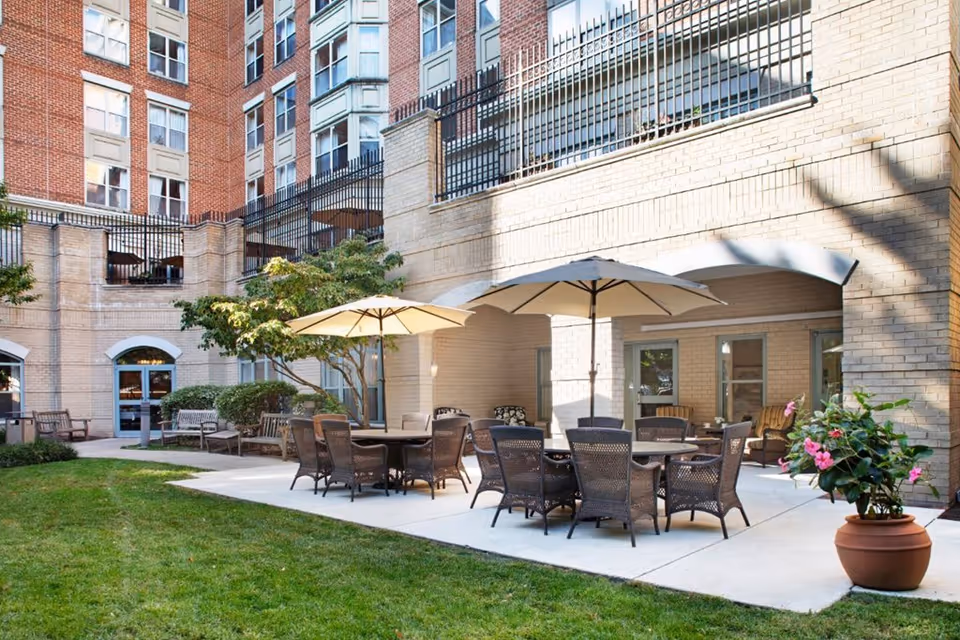 Outdoor patio area at Brighton Gardens of Friendship Heights featuring multiple round tables with umbrellas and wicker chairs on a concrete surface, surrounded by green grass, potted plants, benches, and a multi-story brick building in the background.