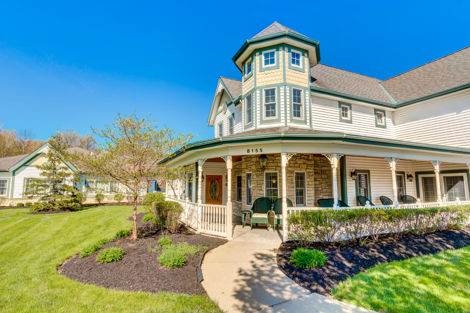 Exterior view of a senior living facility building with white siding, green trim, and a covered porch with green chairs. The building has a turret-like architectural feature and is surrounded by well-maintained landscaping including grass, bushes, and small trees under a clear blue sky.