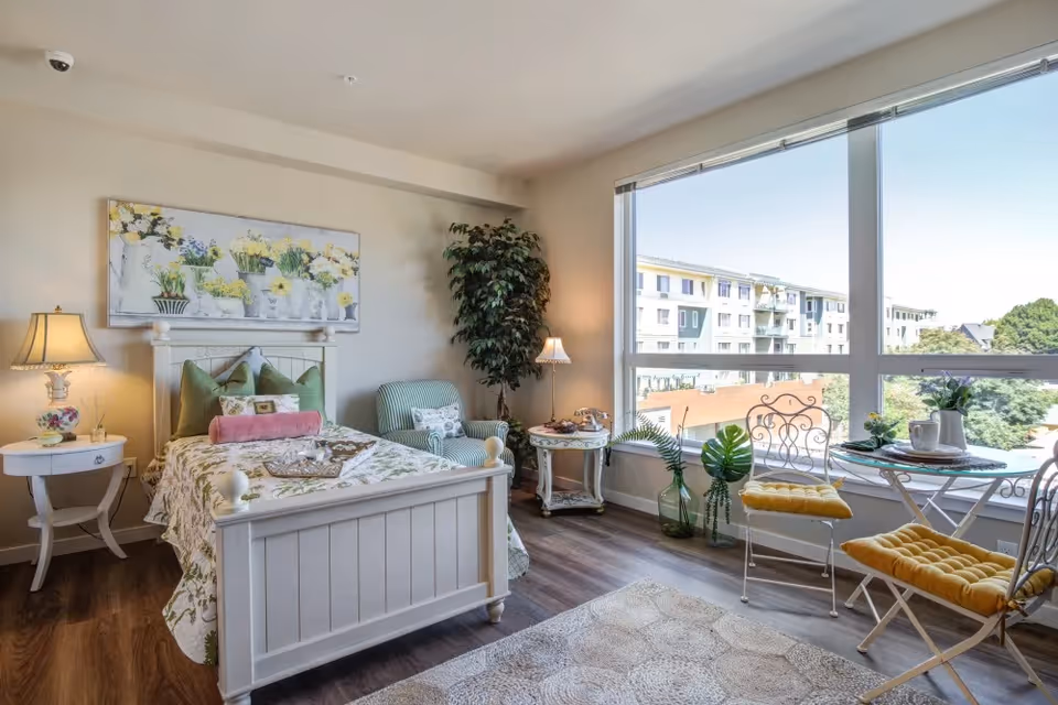 A bright and cozy bedroom with a white wooden bed adorned with green and pink pillows and floral bedding. There is a white nightstand with a decorative lamp on the left side of the bed. A green armchair with a patterned pillow and a small round table with a lamp are placed near a large window. The window offers a view of a multi-story building and greenery outside. Two white metal chairs with yellow cushions and a small round table with a plant and a cup are positioned by the window. The room has wooden flooring and a light-colored rug.