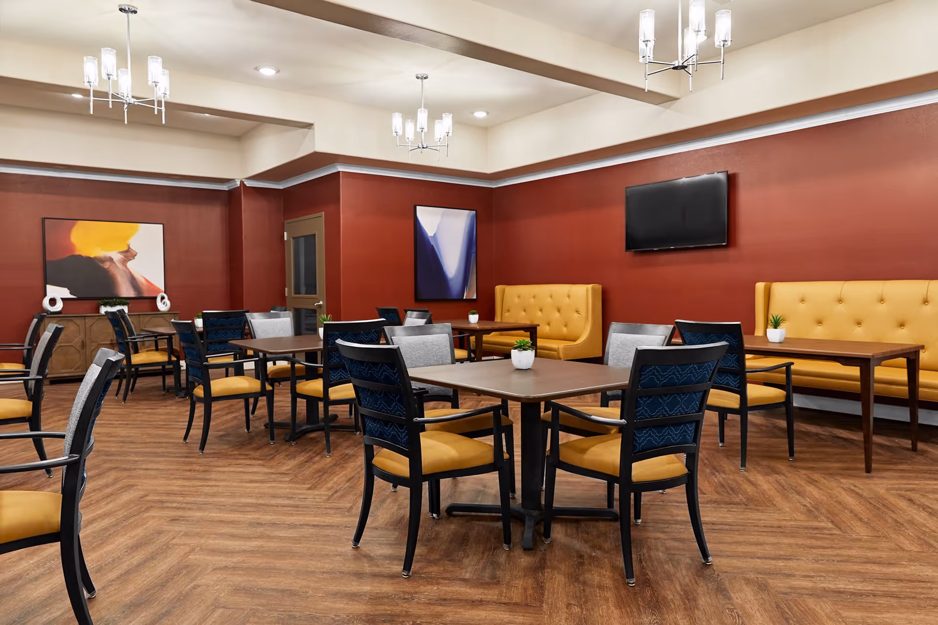 Communal dining room with square tables and chairs, yellow tufted banquettes, wall art and a mounted TV.