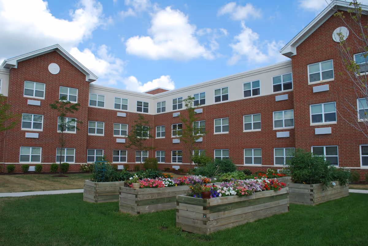 Outdoor garden area with raised wooden flower beds filled with colorful flowers and plants, surrounded by a green lawn. In the background, there is a three-story brick building with many windows under a partly cloudy blue sky.