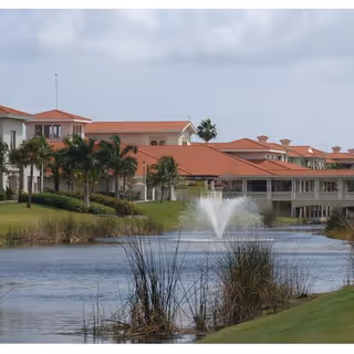 A large senior living complex with red-tiled roofs beside a pond with a central fountain and palm trees.