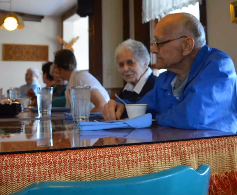 A group of elderly residents sit at a long table in a communal dining room with glasses and bowls.