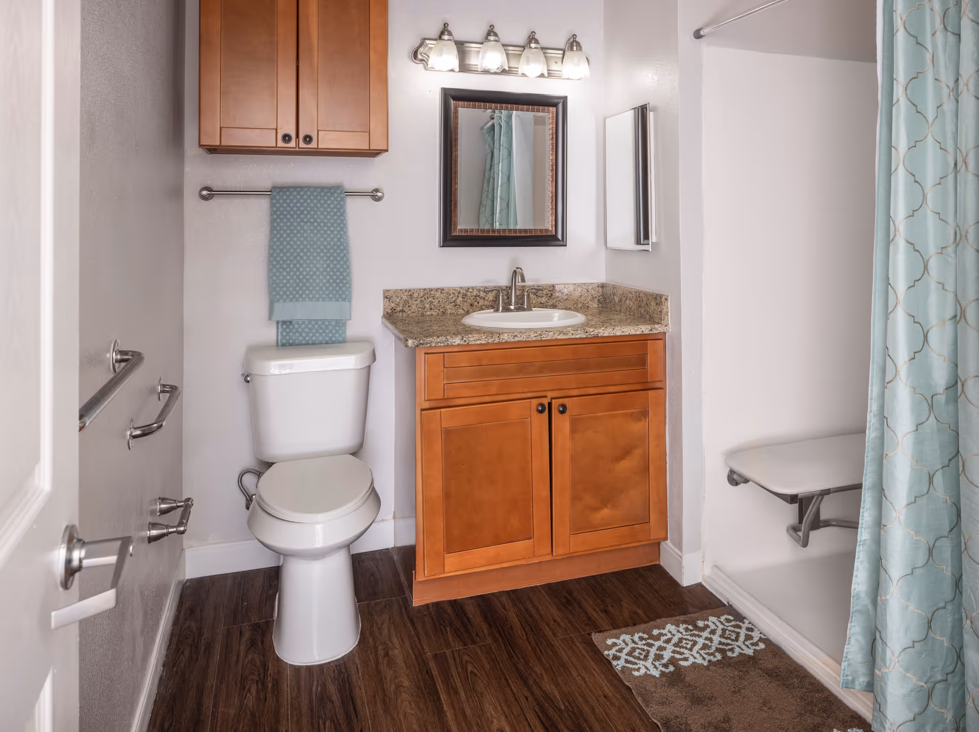 A bathroom with a white toilet, wooden vanity with granite countertop and sink, a mirror above the sink with four light fixtures, a wooden cabinet mounted on the wall, a towel rack with a blue towel, a shower area with a blue patterned curtain and a fold-down shower seat, and a brown and blue patterned bath mat on a dark wood floor.