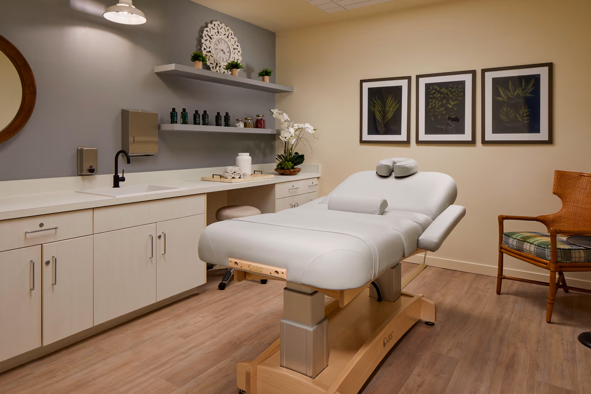 A serene treatment room with a white adjustable massage table, cabinets and countertop, wall shelves and framed botanical prints.
