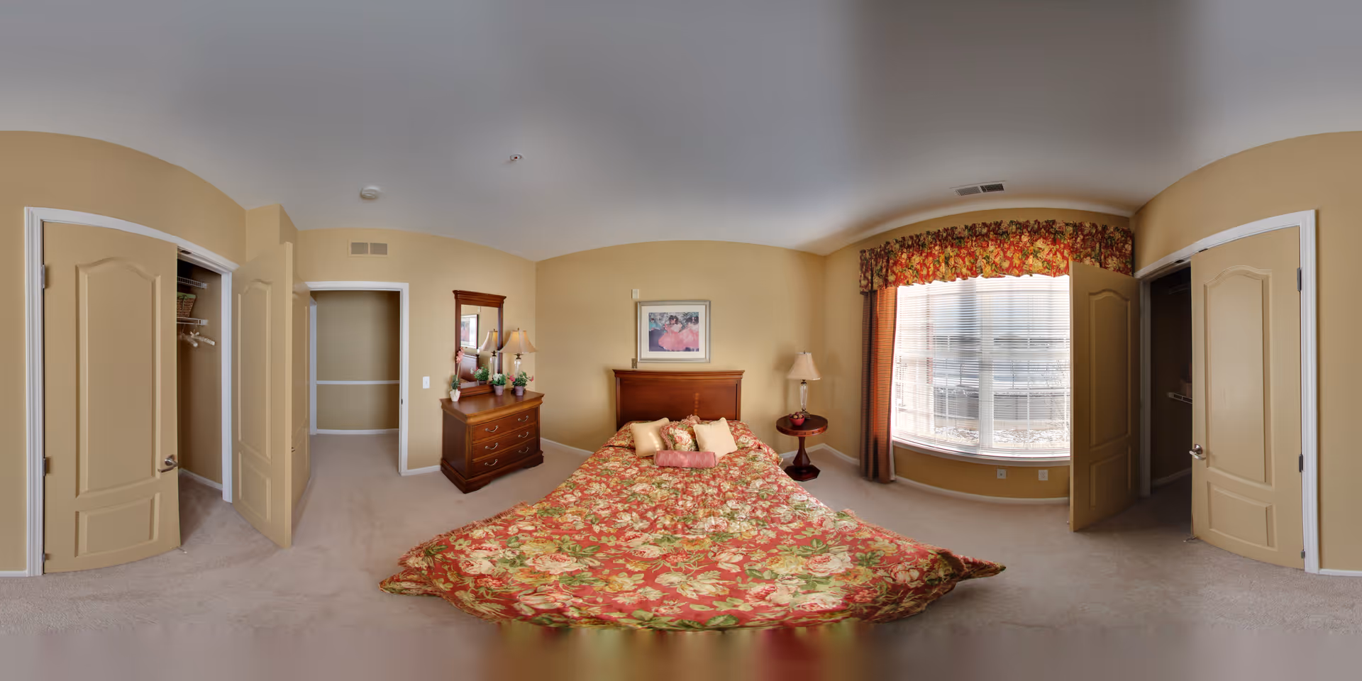 A bedroom with beige walls and carpet, featuring a bed with a floral patterned bedspread in red, green, and beige tones. The bed has several decorative pillows. To the left of the bed is a wooden dresser with a mirror, a lamp, and some decorative items. To the right of the bed is a round wooden side table with a lamp. There is a large window with floral valance and curtains letting in natural light. Two open closets with beige doors are visible on either side of the room.