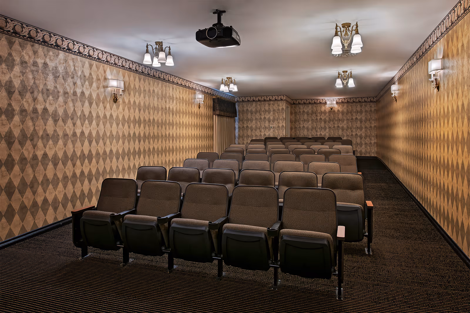 A small theater room with several rows of brown cushioned seats facing a projector mounted on the ceiling. The walls are decorated with a diamond-patterned wallpaper in shades of brown and beige, and there are multiple wall sconces and ceiling lights providing illumination.