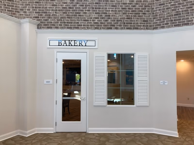 Interior view of a hallway in a senior living facility showing a white door with a glass panel labeled 'Bakery' above it. To the right of the door is a window with white shutters. The walls are light-colored with a brick accent near the ceiling. The floor has a patterned carpet.
