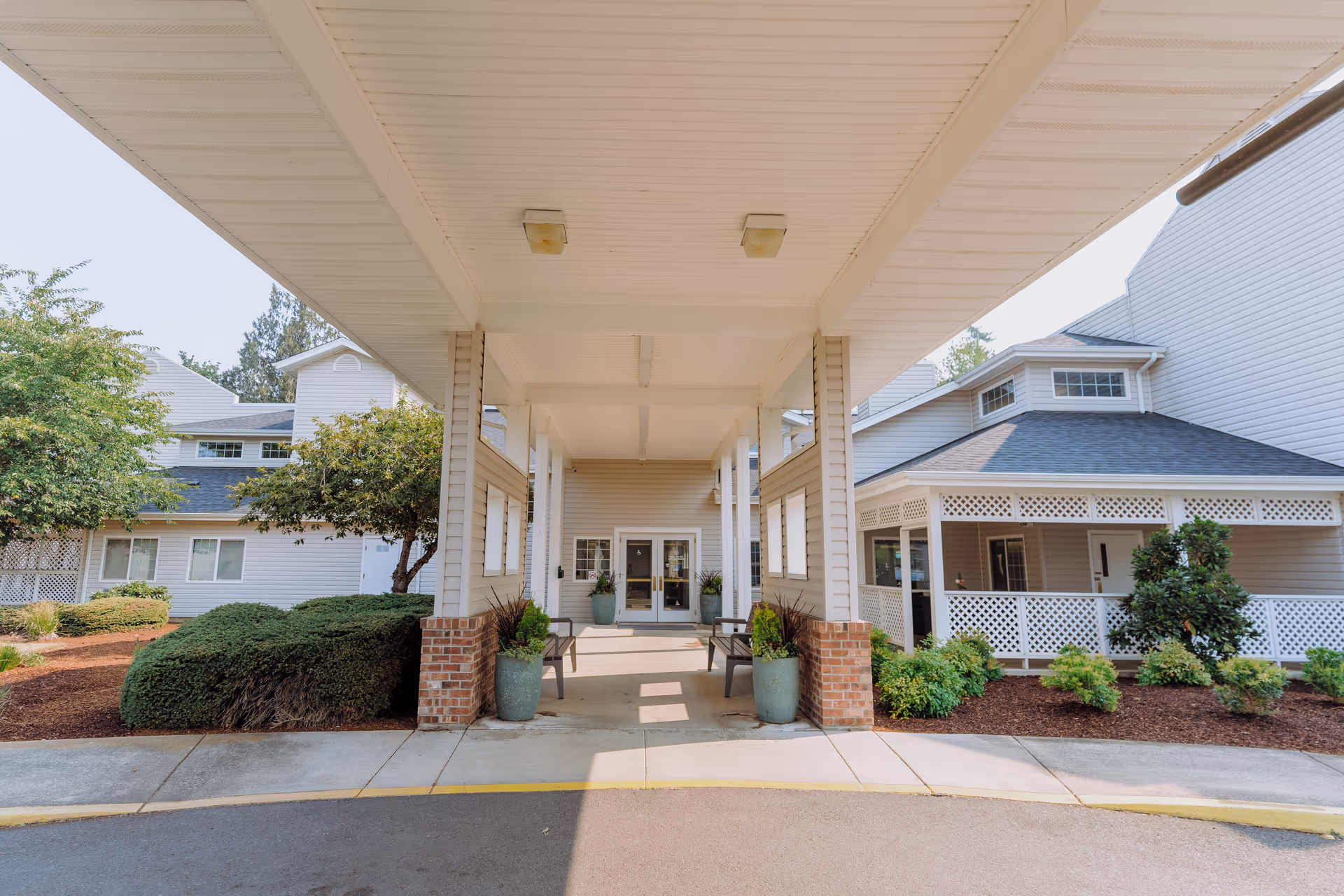 Entrance area of Wiley Creek Senior Living facility showing a covered drop-off zone with benches and potted plants, surrounded by well-maintained bushes and trees, with the building's exterior visible in the background.