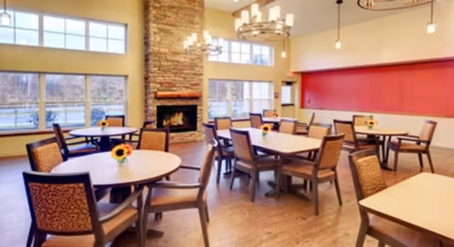 Bright communal dining room with round and rectangular tables, cushioned chairs, a stone fireplace, and large windows.