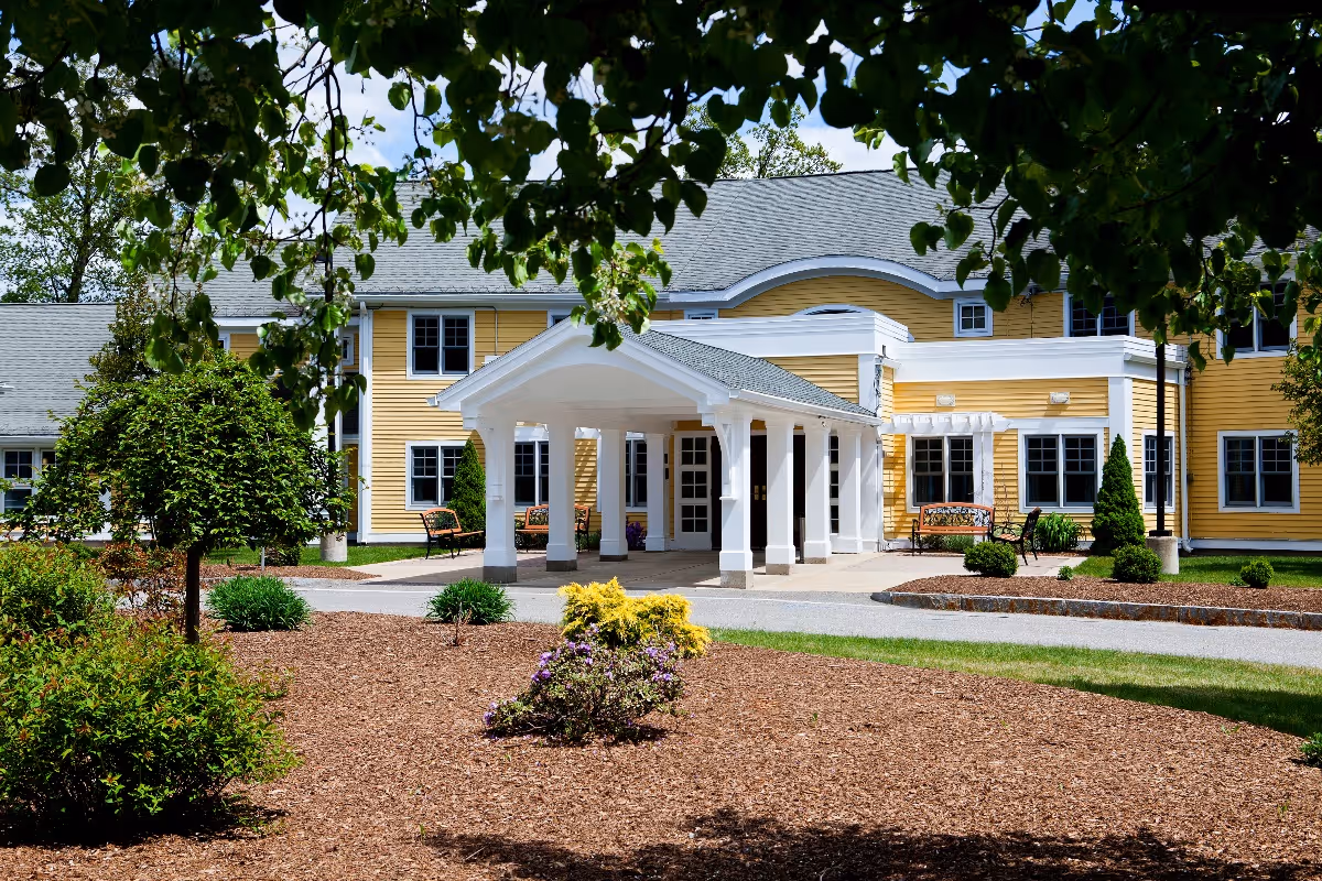 Front exterior view of a yellow senior living facility building with white trim and a covered entrance supported by white columns. The building is surrounded by landscaped grounds with bushes, small trees, and a mulched garden area. There are benches near the entrance and leafy tree branches framing the top of the image.