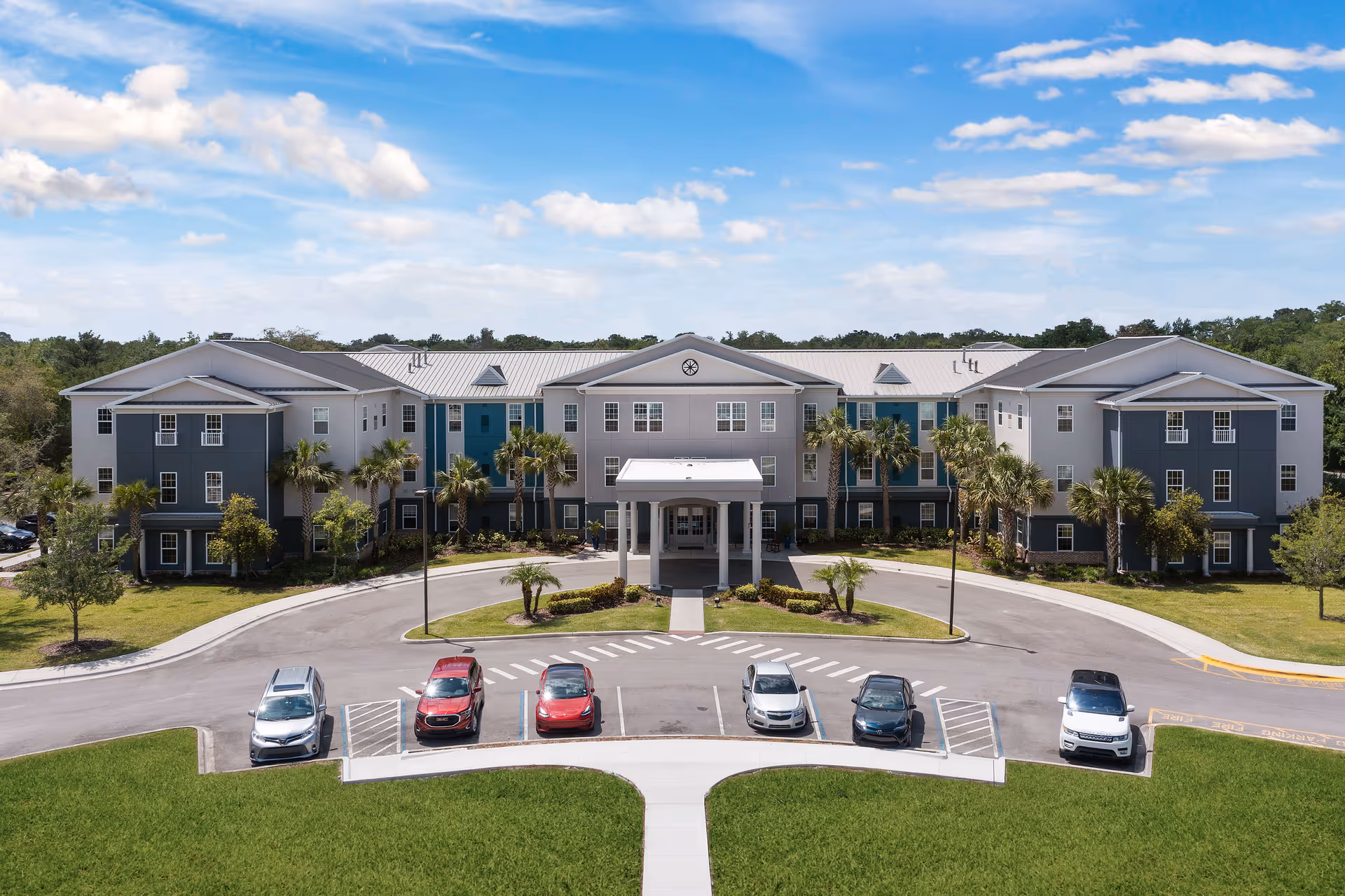 Front exterior view of Gentry Park Orlando, a large three-story building with a covered entrance, surrounded by palm trees and a parking area with several cars. The sky is partly cloudy with blue patches.