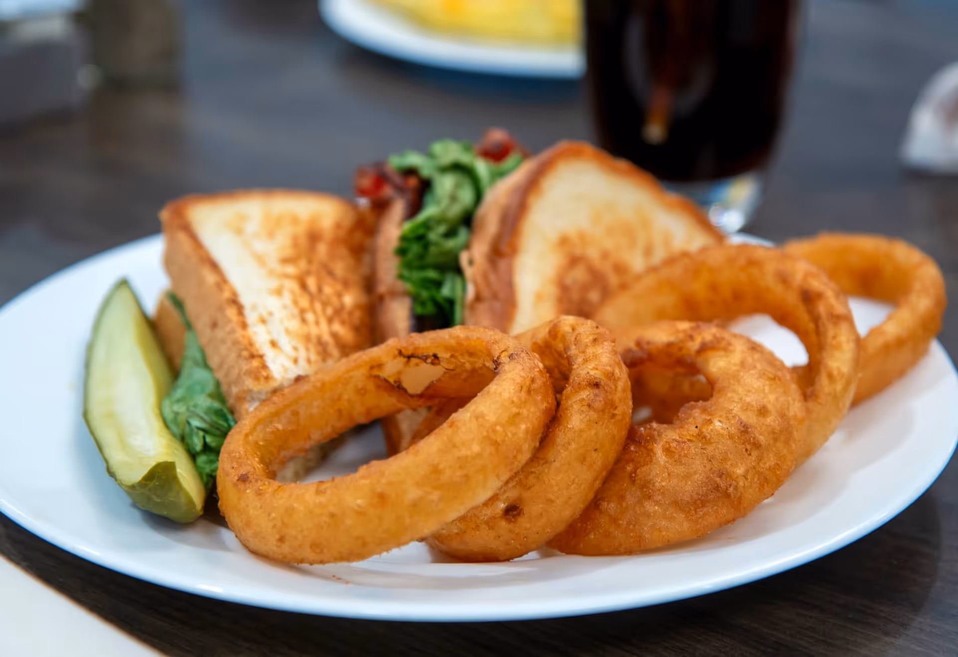 A plate with a sandwich containing lettuce and bacon, a pickle spear, and several golden-brown onion rings, with a glass of dark soda in the background.