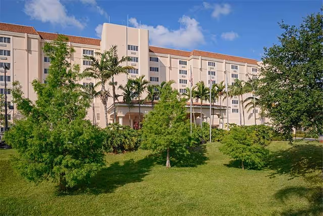 Exterior view of a multi-story senior living facility building with a beige facade and red-tiled roof, surrounded by green trees, palm trees, and a well-maintained lawn under a blue sky with some clouds.