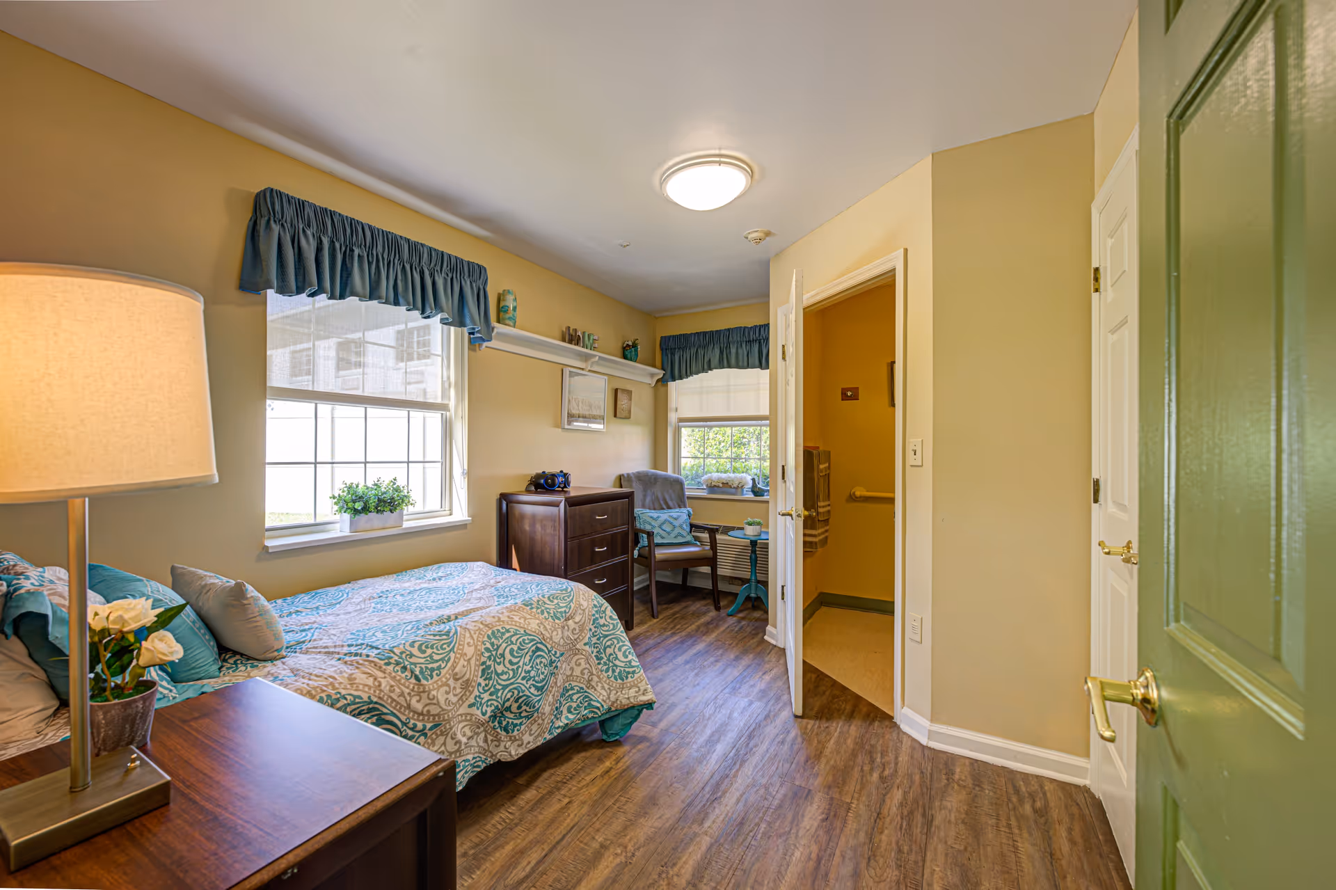 A cozy bedroom in a senior living facility with a single bed covered in a patterned blue and beige quilt. There is a wooden nightstand with a lamp and a small plant, a wooden dresser, and a chair with a small side table near a window with blue valances. The room has wood flooring and beige walls. An open door leads to a bathroom.