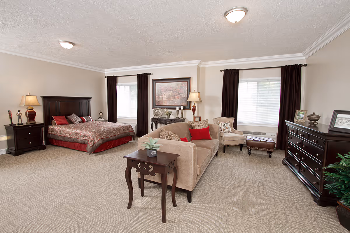 A spacious and well-lit bedroom with a large bed featuring a patterned bedspread and multiple pillows. The room includes a beige sofa with red and beige cushions, a wooden side table with a small plant, a beige armchair, and a dark brown ottoman. There are two windows with dark curtains, a wooden dresser with framed pictures and decorative items, and two lamps providing warm lighting. The walls are light-colored and the floor is carpeted.