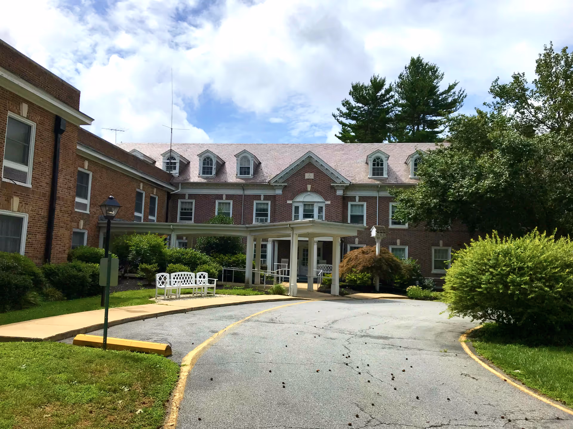 Front exterior view of a large brick building with multiple windows and dormers on the roof. There is a covered entrance with white pillars and a curved driveway leading up to it. The surrounding area has green bushes, trees, and a grassy lawn under a partly cloudy sky.