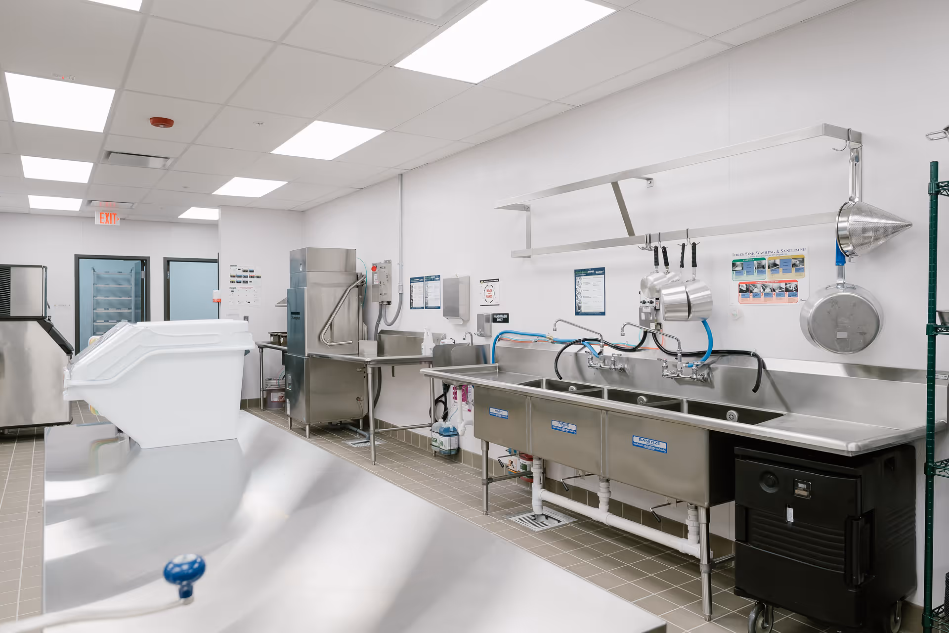 A clean, well-lit commercial kitchen dishwashing area with stainless steel sinks, hanging pots and pans, a large dishwasher, and various cleaning supplies. The room has white walls, a tiled floor, and fluorescent ceiling lights.
