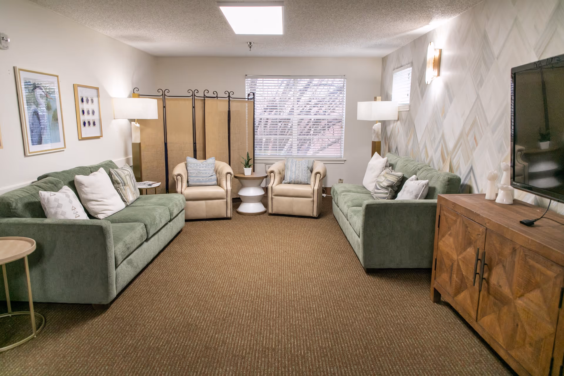A cozy living room with two green sofas facing each other, two beige armchairs with blue striped pillows, a wooden cabinet with a TV on top, two floor lamps, a small round side table, and a window with blinds letting in natural light. The walls are decorated with framed artwork and a patterned wallpaper on one side.