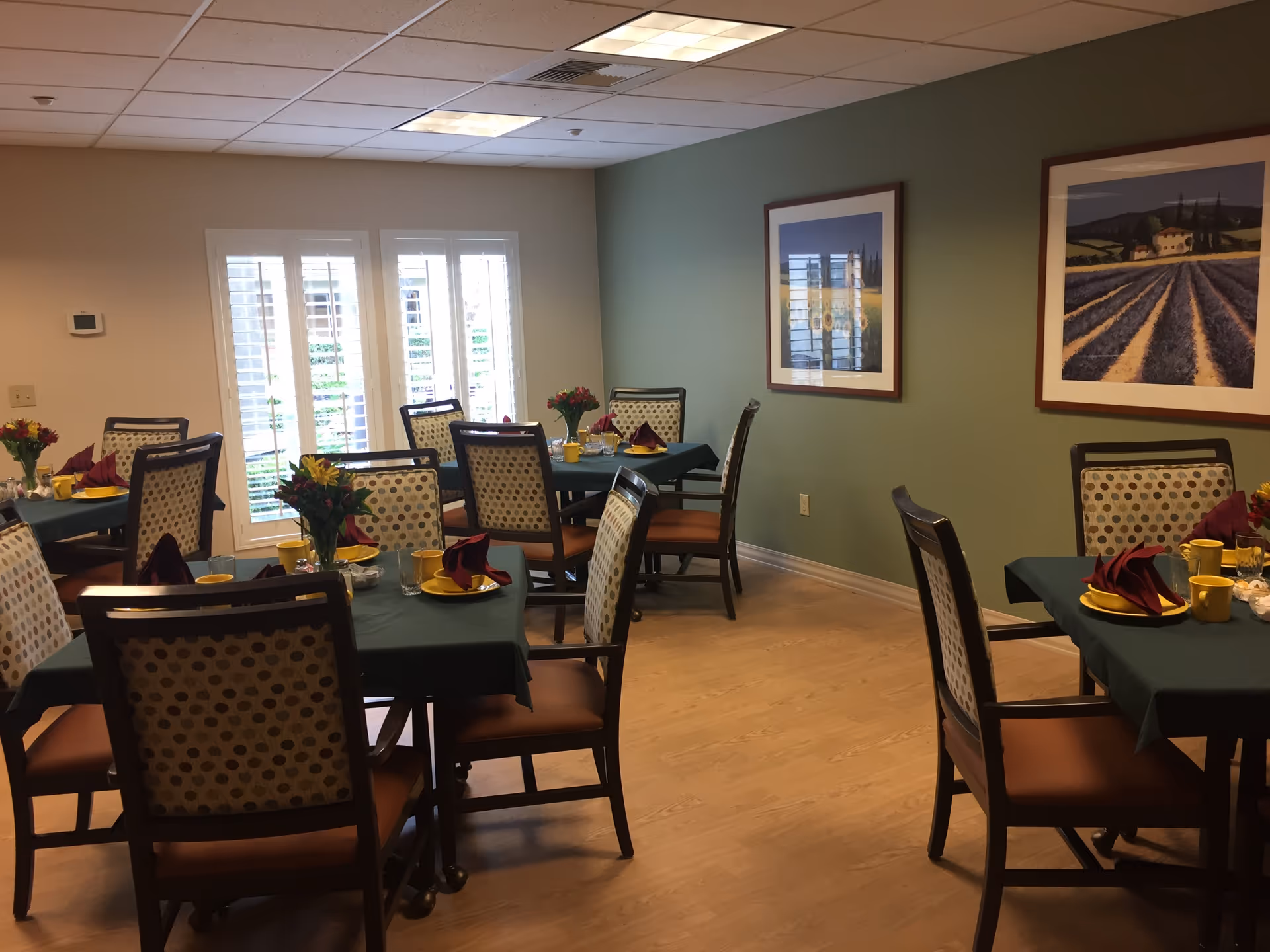 Dining room with several tables covered in dark green tablecloths, each set with yellow plates, cups, glasses, and burgundy folded napkins. Chairs with patterned backs and orange seats surround the tables. The room has light wood flooring, a green accent wall with two framed landscape paintings, and windows with white shutters letting in natural light.