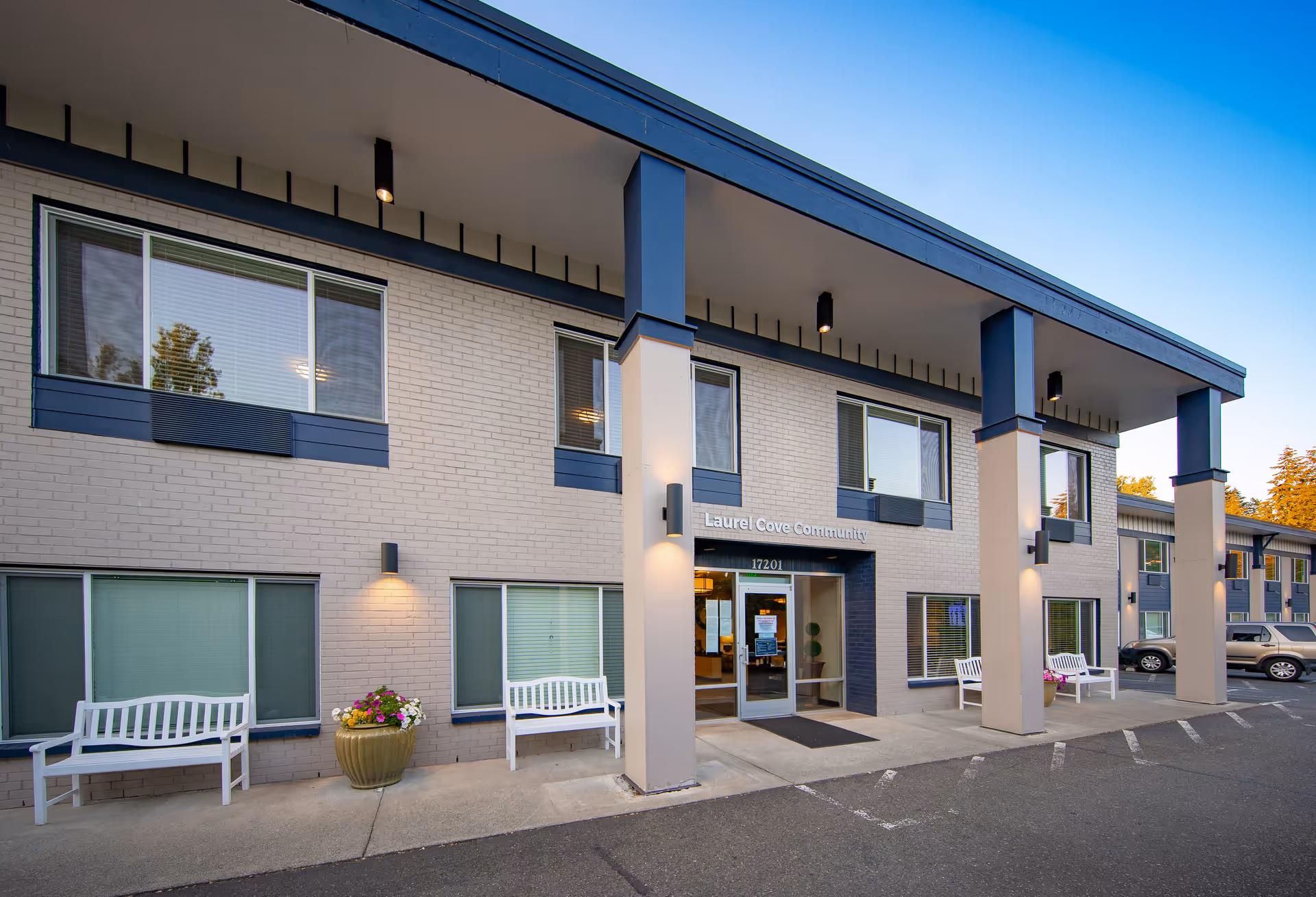Exterior front view of Laurel Cove Community assisted living and senior care facility with white benches, flower pots, large pillars, and multiple windows under a clear blue sky.