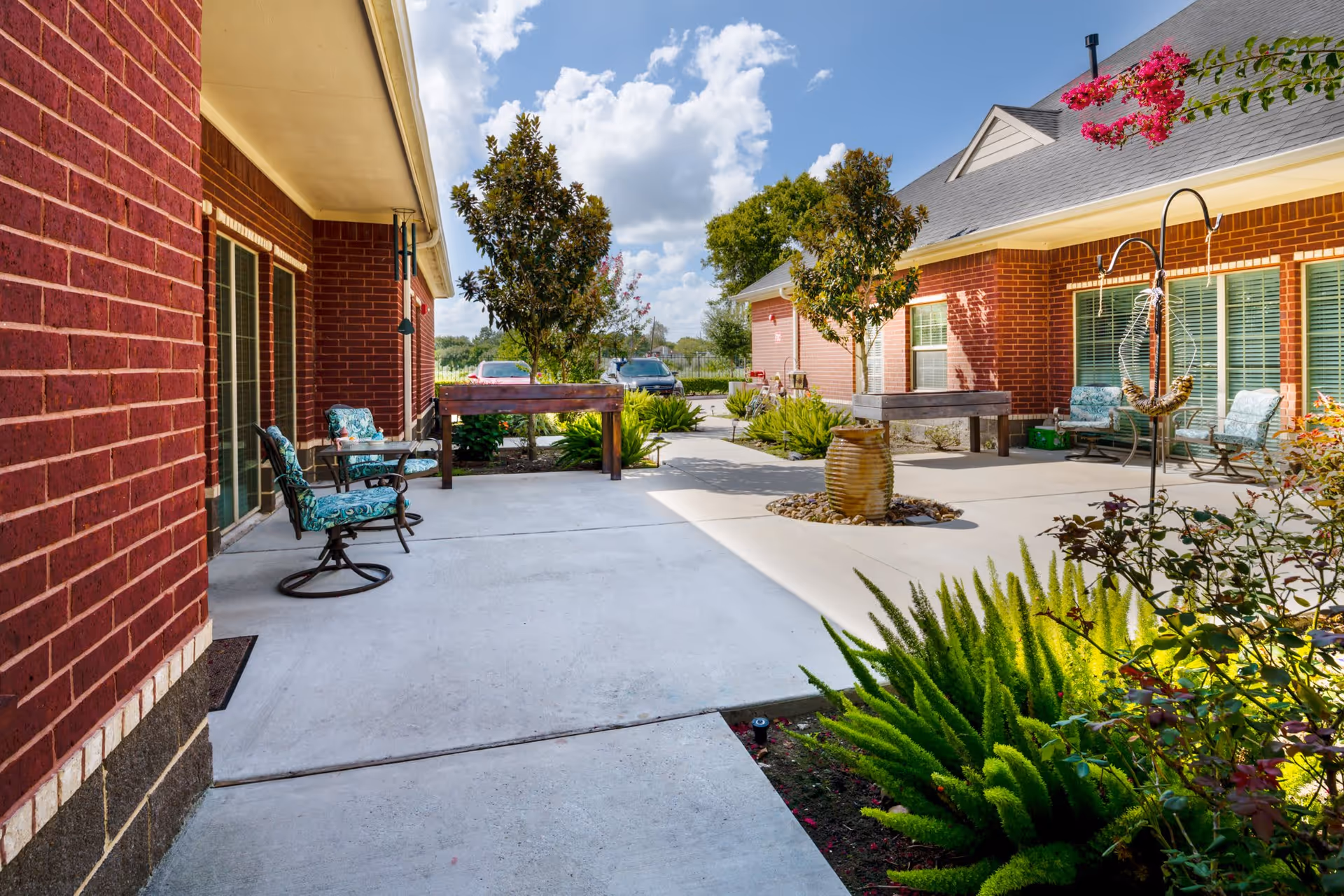 Outdoor patio area between two red brick buildings with concrete walkways, green plants, trees, and several cushioned chairs arranged for seating. A large ceramic pot is placed in the center of the patio, and the sky is partly cloudy.