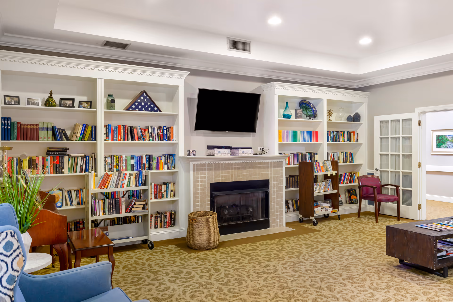 A cozy living room area in Brookdale Meadowmont featuring built-in white bookshelves filled with books and decorative items, a wall-mounted flat screen TV above a tiled fireplace, a wicker basket on the floor, a blue armchair with a patterned pillow, a wooden side table, and a maroon chair near a glass-paneled door. The room has beige patterned carpet and recessed ceiling lights.