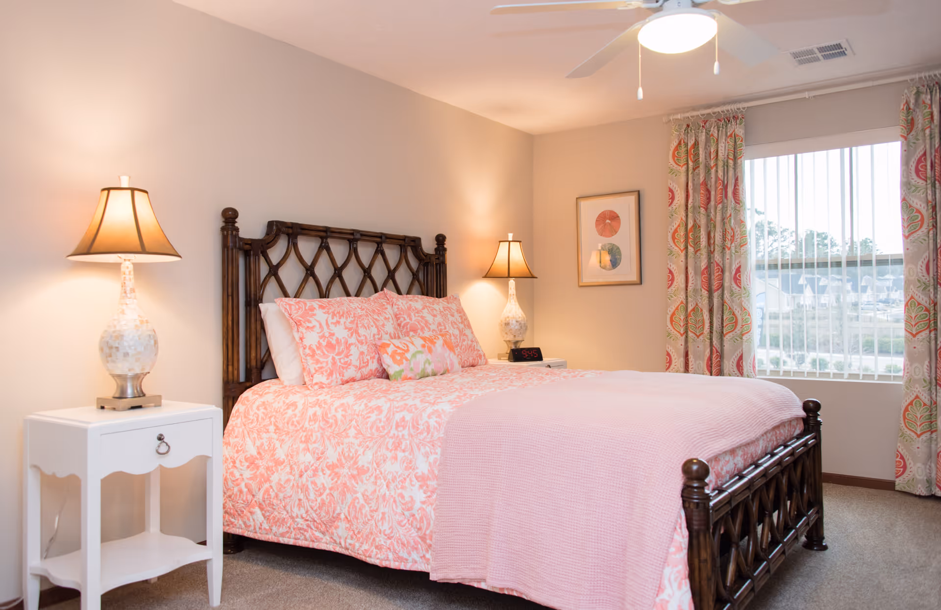 Bright bedroom with a dark wooden bedframe dressed in pink patterned bedding, two bedside lamps, a white nightstand, and a window with patterned curtains.