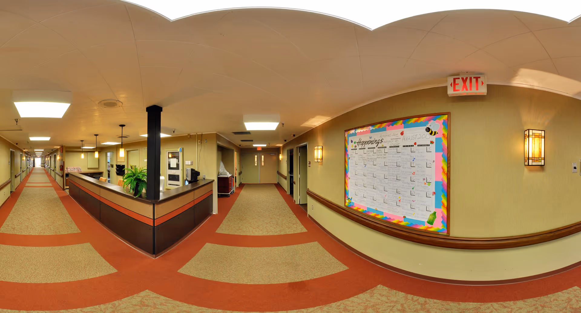 A wide hallway in a senior living facility with a reception desk on the left side and a bulletin board displaying a calendar of happenings on the right wall. The hallway is carpeted with a beige and red pattern, has beige walls, ceiling lights, and handrails along the walls. An exit sign is visible above a door at the end of the hallway.