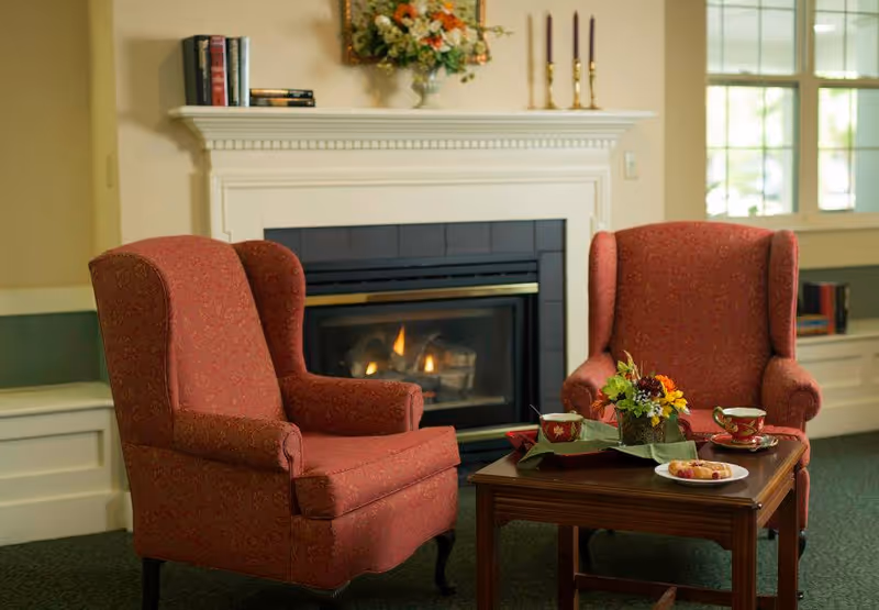 Cozy seating area with two red upholstered armchairs facing a lit fireplace and a small table set with tea and flowers.